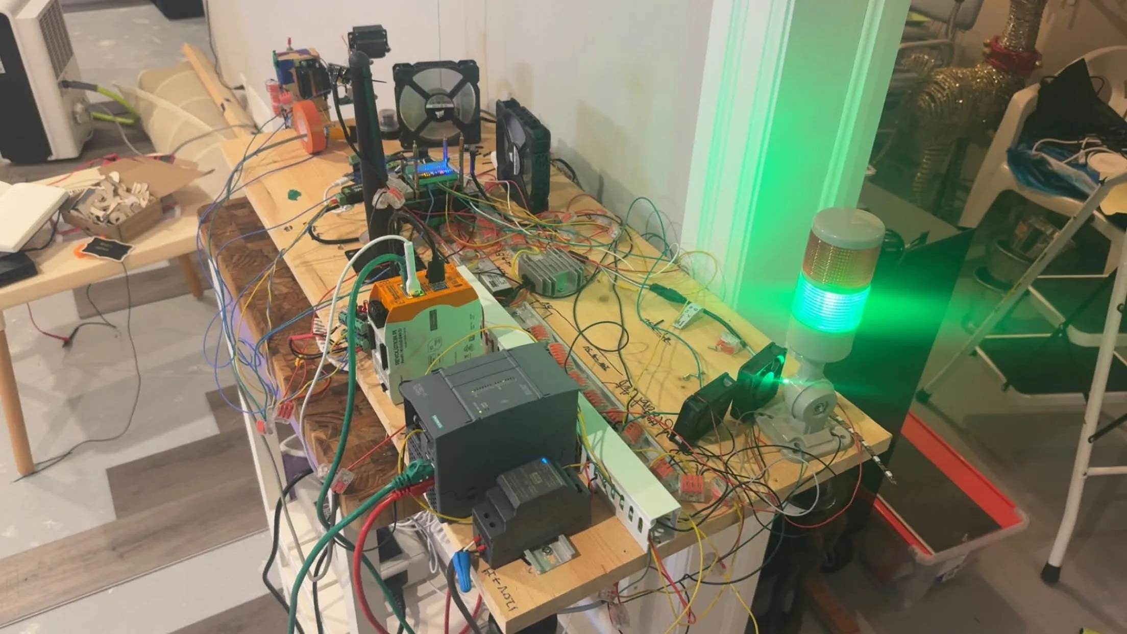 The Harvester testbed on a workbench: Siemens S7-1200 PLC, orange Kunbus RevPi, relay banks, fans, and a green industrial stack light.